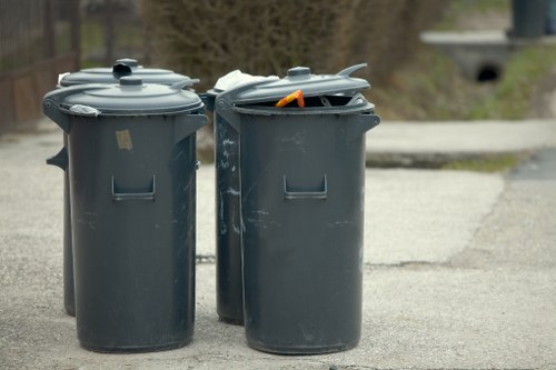 Two dark grey plastic wheelie bins with closed lids are positioned on a paved driveway, with one bin having a partially open lid revealing an orange-handled tool or object inside. The bins are slightly scuffed and weathered, indicating regular use, and sit in an outdoor residential or semi-urban environment. The background shows blurred natural elements like trees and shrubbery, with a low curb and a small concrete bridge or culvert visible further back. The scene captures an everyday rubbish collection setting, aligning with rubbish removal services in the Colindale area, part of the London borough of Barnet, known for residential properties and local waste disposal needs. Office Clearance Colindale's services might include the collection and disposal of household waste from such areas, ensuring proper rubbish removal and waste management.