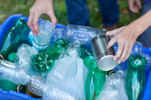 Close-up view of a person's hands sorting plastic waste at a recycling collection, with one hand holding a clear plastic water bottle and the other placing a metal can into the bin. The collection container is filled with various recyclable plastics, including bottles of different sizes and colors, primarily clear and green. The background shows a grassy outdoor area, indicative of a community clean-up or recycling event, possibly in or near Colindale. The scene emphasizes the importance of proper rubbish separation, aligned with services offered by Office Clearance Colindale, which specialises in waste management and rubbish removal in the local area. The image highlights the textures of the plastic bottles and metallic cans, with the bottles having a glossy, smooth surface and the can featuring a shiny, reflective finish. Overall, the visual captures a moment of active waste sorting within an outdoor setting dedicated to environmental responsibility.