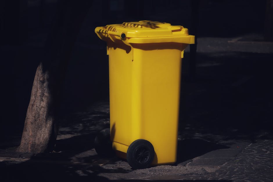 A yellow plastic wheeled rubbish bin with a closed lid, positioned on a dark pavement surface beside a weathered concrete curb. The bin's smooth, glossy surface reflects minimal light and appears to b