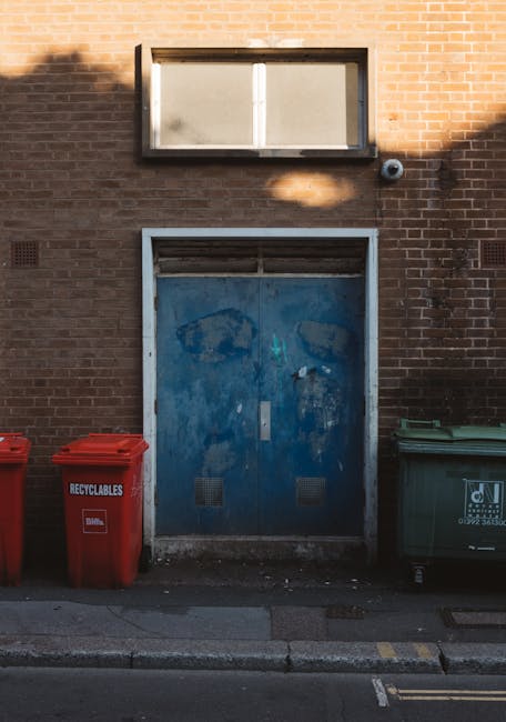 A yellow plastic wheeled rubbish bin with a closed lid, positioned on a dark pavement surface beside a weathered concrete curb. The bin's smooth, glossy surface reflects minimal light and appears to b