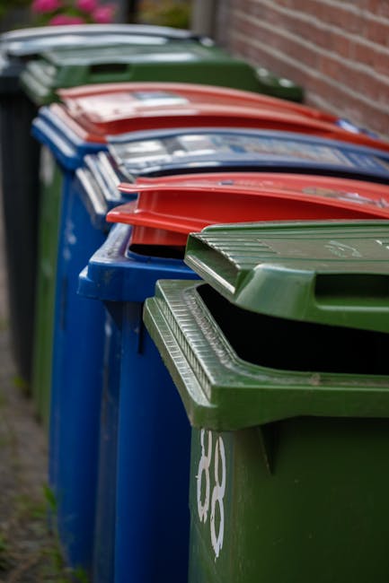 A black wheeled rubbish bin labeled 'St. John's' positioned on a paved sidewalk at night, filled with various discarded items including cardboard boxes, plastic containers, and paper waste, some of wh