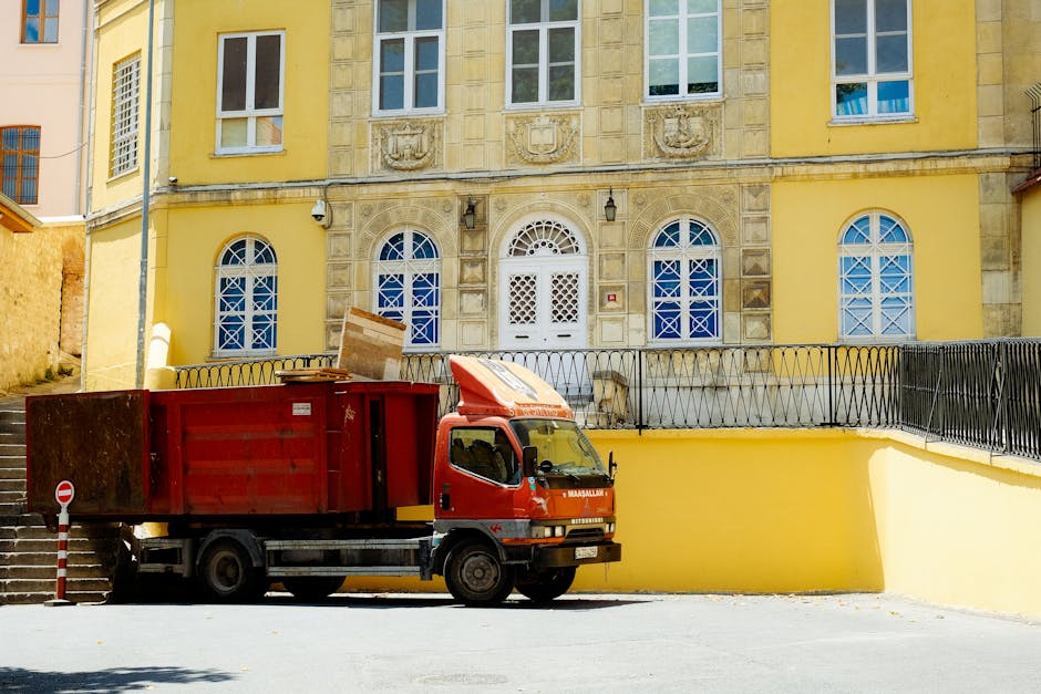 A small, dark red flatbed truck parked on an urban street in front of a modern building with large glass windows and white concrete panels. The truck's cargo area is filled with various waste material